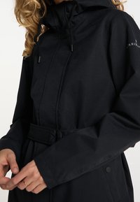 Veste noire imperméable avec un col haut, une capuche à cordon, une taille ceinturée et une texture lisse. Comprend des poches latérales et des accents à boutons.