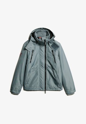 Giacca isolata grigia chiara con colletto alto, tasche con zip e una patch sulla spalla sinistra. Presenta un tessuto testurizzato e un cappuccio.