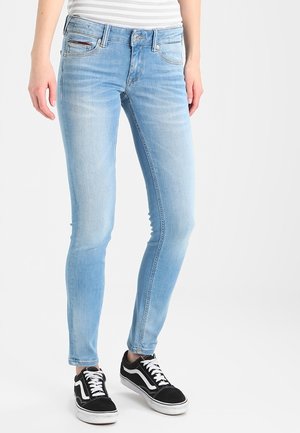 Personne portant un jean skinny bleu clair et des baskets noires avec des lacets blancs, debout devant un fond blanc uni.