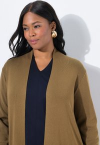 Frau mit langen schwarzen Haaren, die ein dunkelblaues Oberteil, eine braune Strickjacke und große goldene Ohrringe trägt und vor einer weißen Wand steht.