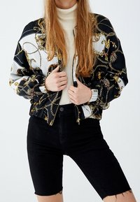 Veste bomber noire avec un motif de chaînes dorées, fermeture éclair et poignets côtelés. Portée avec un col roulé crème et un short en denim noir.