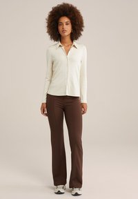 Camisa de botones de manga larga color crema con rayas verticales, combinada con pantalones acampanados marrones y zapatillas gris, de pie contra un fondo neutro.