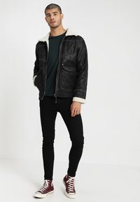 Schwarze Lederjacke mit einem weißen Lammfellkragen, Reißverschluss und vier Taschen, getragen über einem dunkelgrünen Oberteil und schwarzen Skinny Jeans.