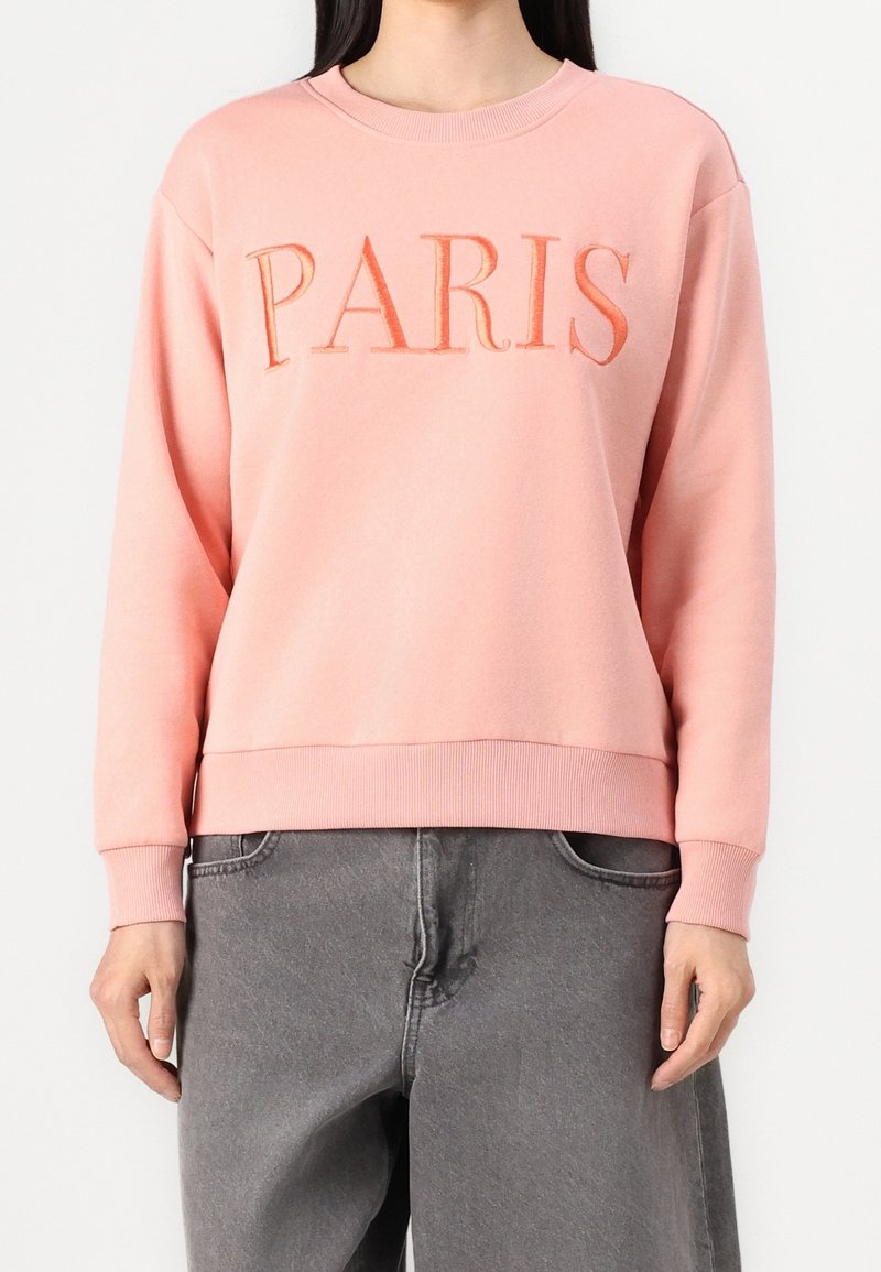 Femme portant un sweat-shirt rose avec "PARIS" brodé en orange et un pantalon gris ample, debout sur un fond blanc.