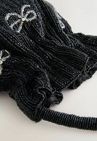 Melnā, krellītēm rotāta clutch somiņa ar sakumpušo dizainu. Iezīmējas ar spīdīgiem sudrabainiem akcentiem, kas atgādina loku, un teksturētu, austu siksniņu.