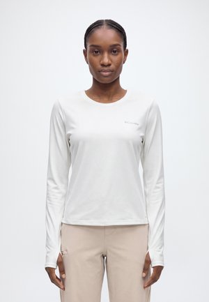 Une femme présente une chemise Columbia blanche à manches longues et un pantalon beige sur un fond uni et clair.
