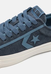 Zapatilla deportiva de ante gris con acentos azules, con cordones planos, suela de goma y un logo de estrella en el lateral. Diseño clásico de corte bajo.