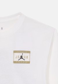 Vit bomullströja med en rektangulär etikett som har "AUTHENTIC JORDAN BRAND," guldfinish och en svart jumpman-logotyp.