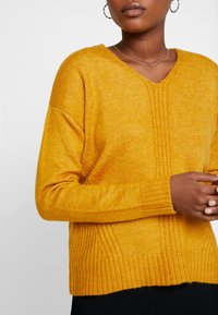 Mosterdgele V-hals trui met geribbelde textuur, verlaagde schouders en zijpaneel details. Zachte stof, losse pasvorm en lange mouwen.