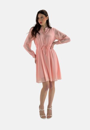 Abito lungo a maniche lunghe di colore rosa chiaro, con apertura frontale a bottoni, vita cinchata e gonna fluente. Abbinato a sandali altissimi bianchi con cinturino.