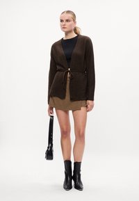 Cardigan de punto marrón con cierre de lazo, combinado con una falda de cuadros, top negro y botines negros de tacón, llevando un pequeño bolso de hombro.