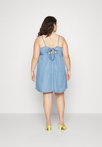 Vestido de ganga com alças spaghetti ajustáveis, fecho nas costas em forma de buraco e um detalhe amarrado. A modelo usa sandálias de salto amarelo brilhante.