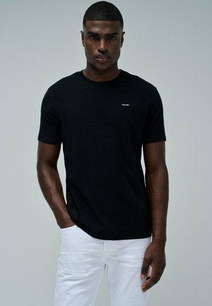 Camiseta negra de algodón con mangas cortas, que presenta una pequeña etiqueta de la marca en el lado izquierdo del pecho. El modelo lleva pantalones blancos y tiene una expresión neutral.