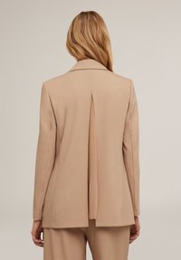 Beige bleiseri, jossa on keskipitkä istukku takana, standardimittainen kaulus, muotoutunut istuvuus ja jämäkät olkapäät, valmistettu sileästä kankaasta.