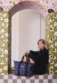 Kvinna med blont hår i mörka kläder som sitter under en blommig båge och håller en stor marinblå tygväska med blommönster på ett rutigt golv.