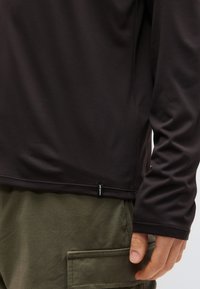 Κοντινό πλάνο ενός ατόμου που φοράει μαύρο μακρυμάνικο μπλουζάκι με μικρή ετικέτα "Patagonia" και ελιάς-πράσινο παντελόνι με μπροστινή τσέπη.