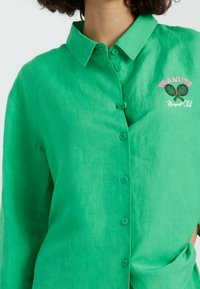 Groene linnen blouse met een knoopsluiting aan de voorkant, voorzien van een kraag en een geborduurd "Peanuts"-logo met tennisrackets aan de linkerkant.