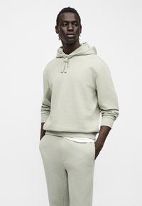 Νέος άνδρας με σκουρόχρωμο δέρμα που φορά ανοιχτό γκρι φούτερ με κουκούλα και αντίστοιχο παντελόνι φόρμας, τα χέρια στις τσέπες, κοιτάζει πλάγια σε απλό φόντο.