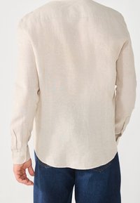 Beige linnen overhemd met lange mouwen, een afgeronde zoom en knoopmanschetten, gedragen over blauwe spijkerbroek. Eenvoudig design, lichtgewicht textuur.