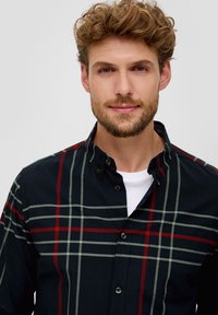 Zwarte en groene geblokte button-up shirt met rode accenten; heeft een kraag, knoopsluitingen en een textuur stof; gelaagd over een wit T-shirt.