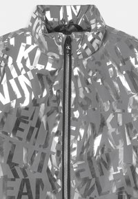 Veste argentée réfléchissante avec un motif de texte audacieux sur toute sa surface. Dotée d'un col haut et d'une fermeture éclair noire à l'avant. Texture lisse et brillante.