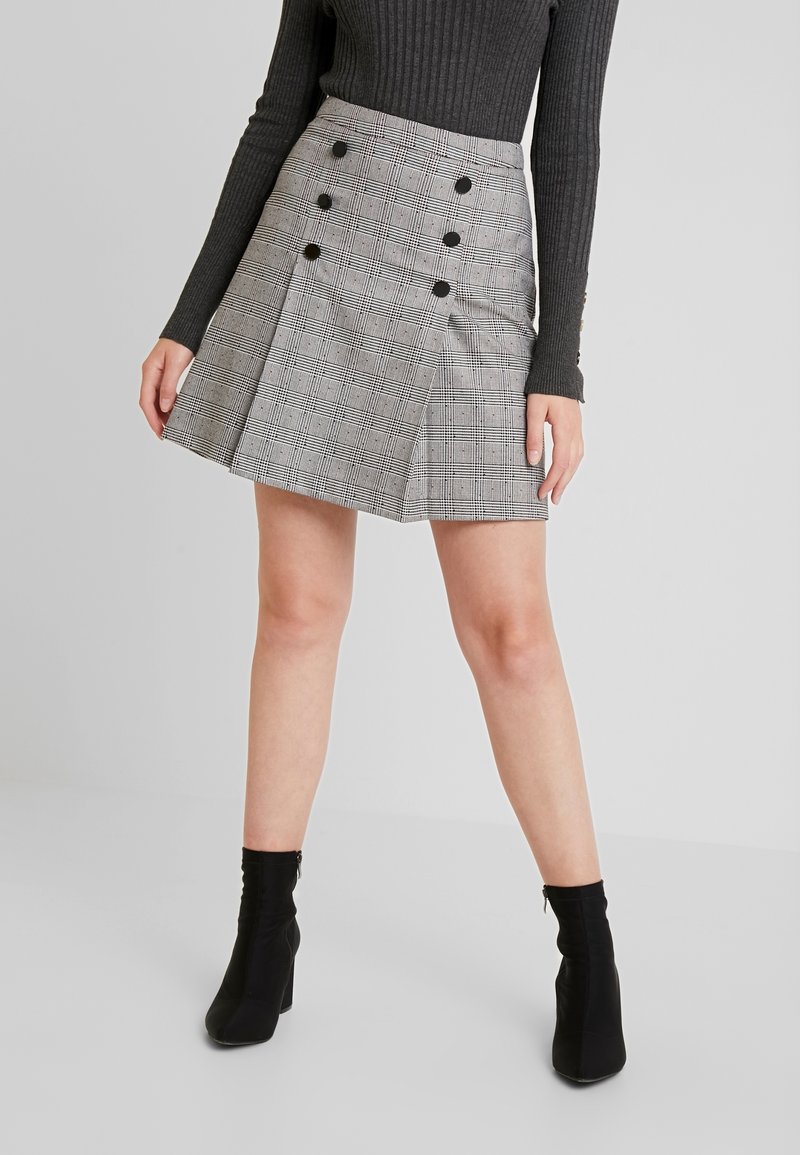 Femme portant une mini-jupe à carreaux grise avec des boutons noirs, associée à un haut noir côtelé à manches longues et des bottines noires.
