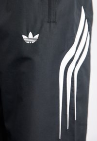 Μαύρες αθλητικές σορτς με λευκό λογότυπο Adidas και τρεις κατακόρυφες λευκές ρίγες στο πλάι, φτιαγμένες από λείο, ελαφρύ ύφασμα.