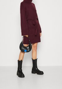 Burgundy stickad klänning med midjebälte, långa ärmar och hög krage. Matchas med svarta klumpiga klackstövlar och en liten svart handväska.