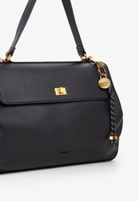 Bolso de mano de cuero negro con un acabado texturizado, herrajes dorados, cierre de solapa y un charm decorativo con una correa trenzada.