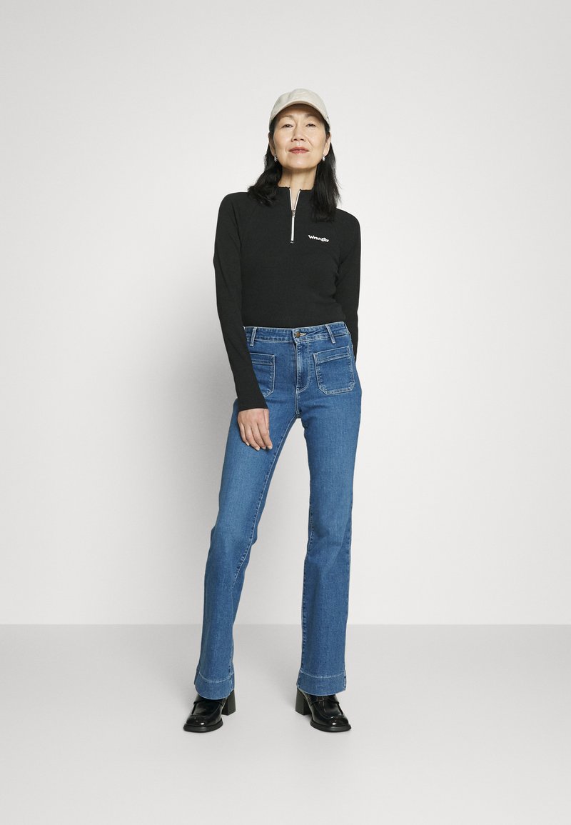 Schwarzes Langarmoberteil mit einem Halb-Reißverschluss-Kragen, kombiniert mit blauen Schlagjeans und schwarzen Stiefeletten mit Absatz, accessoriert mit einer hellen Kappe.