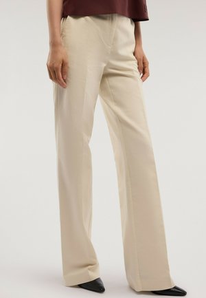 Femme portant un pantalon large beige, des chaussures noires pointues et un haut bordeaux sur un fond clair uni.