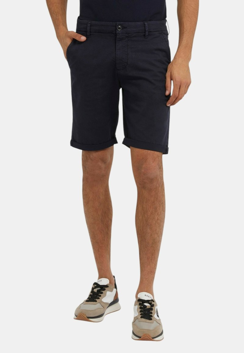 Pantaloni corti in cotone blu navy con risvolti, tasche laterali e chiusura con bottone. Abbinati a scarpe da ginnastica beige e grigie con lacci neri.