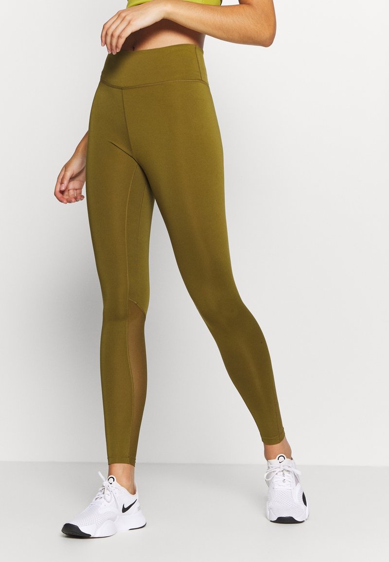 Oliwkowe legginsy z wysokim stanem o gładkiej teksturze, z siateczkowymi panelami po bokach i wzmocnionymi szwami. W połączeniu z białymi butami sportowymi.