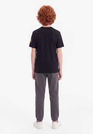 Kind met krullend rood haar staat met de rug naar de kijker, draagt een zwart T-shirt, grijze broek en witte sneakers op een witte achtergrond.