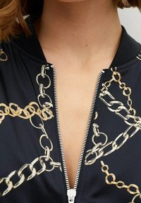 Chaqueta bomber negra con cierre de cremallera en la parte delantera, adornada con un patrón de cadena dorada y un cuello acanalado. Tejido suave con un silueta ajustada.