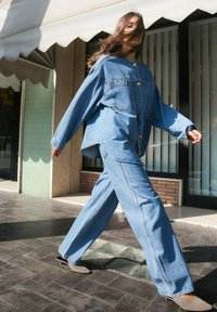 Tenue en denim avec une veste bleue surdimensionnée et un jean à jambe large. Texture lisse avec des détails de couture visibles. Des chaussures grises complètent le look.