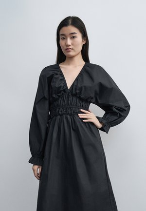 Kvinne iført svart kjole med lange ermer, dyp V-hals og innsvingt midje, står med den ene hånden på hoften mot ensfarget grå bakgrunn.