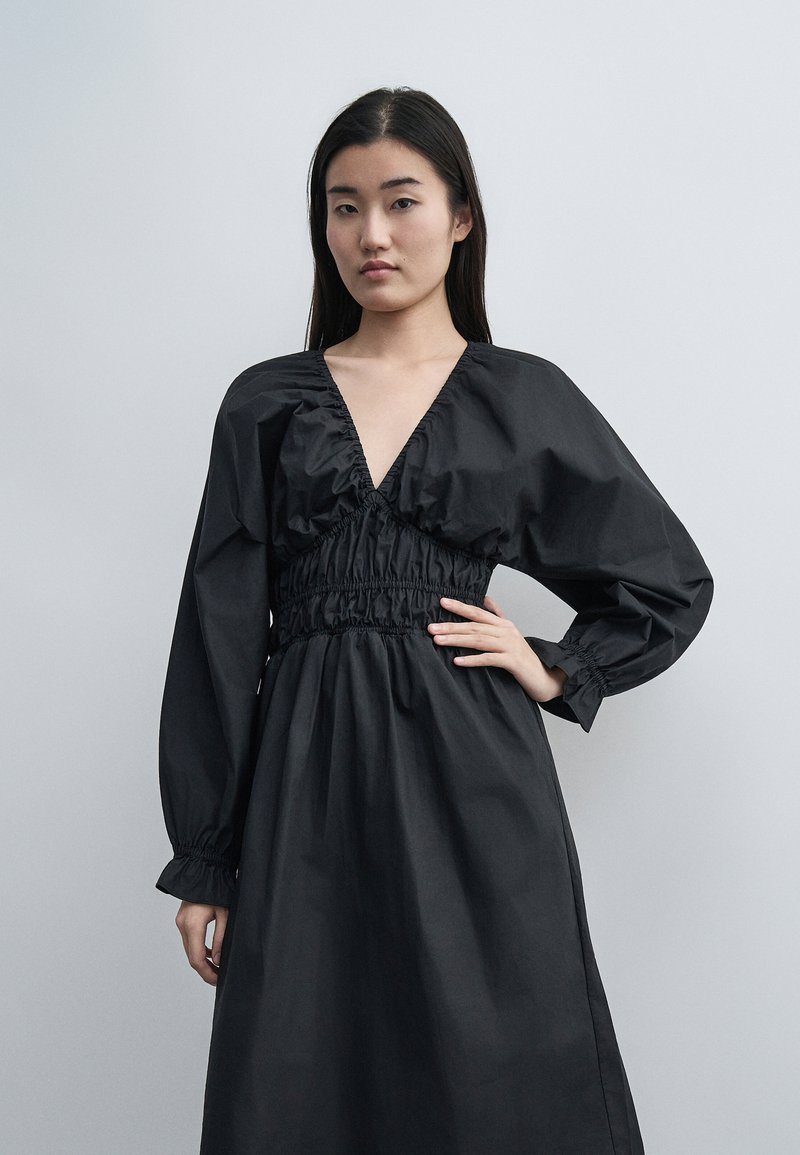 Γυναίκα που φοράει μαύρο φόρεμα με μακριά μανίκια, βαθύ V-λαιμόκοψη και ζωνάκι στη μέση, στέκεται με το ένα χέρι στη μέση σε απλό γκρι φόντο.