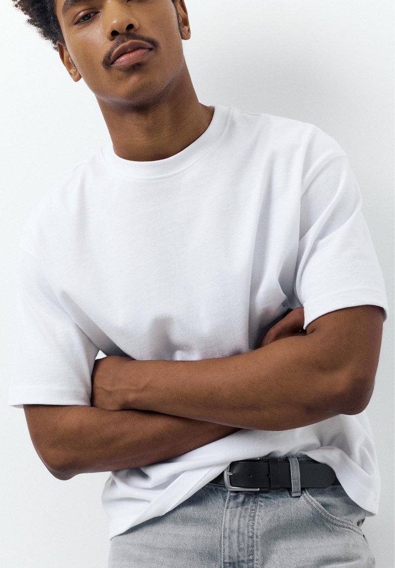 Jeune homme aux bras croisés portant un t-shirt blanc et un pantalon gris clair avec une ceinture noire, sur fond blanc.