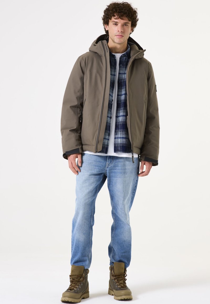 Chaqueta marrón, aislante y con capucha, superpuesta a una camisa de cuadros azul y una camiseta blanca. Unos vaqueros azul claro y botas de senderismo marrones completan el conjunto.