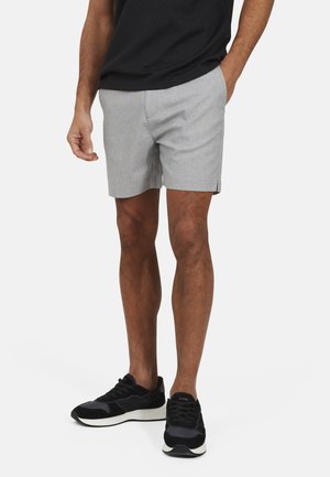 Uomo con pantaloncini grigio chiaro, sneakers nere e maglietta nera, in piedi con una mano in tasca su sfondo bianco.