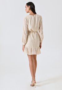 Cremefarbenes Kleid mit Zickzackmuster, langen Ärmeln und Rüschensaum. Der Stoff wirkt strukturiert. Das Model trägt Riemchenabsätze.
