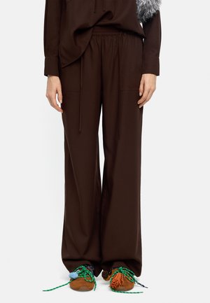 Osoba ubrana w luźne, ciemnobrązowe spodnie i brązowe tenisówki z kolorowymi sznurówkami oraz ozdobami z frędzli, stojąca na jednolitym tle.