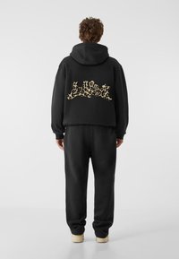 Fekete kapucnis pulóver, rövidített kialakítással, leopárdmintás grafikával a háton. Megfelelő fekete melegítőnadrággal és világos színű cipőkkel párosítva.