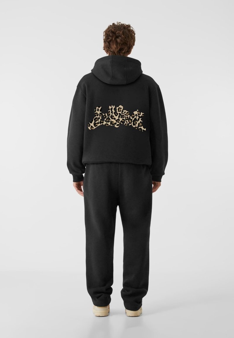 Fekete kapucnis pulóver, rövidített kialakítással, leopárdmintás grafikával a háton. Megfelelő fekete melegítőnadrággal és világos színű cipőkkel párosítva.