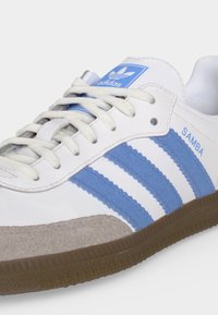 Chaussure blanche Adidas Samba avec des rayures bleu clair, semelle en gomme, bout en daim beige et détails de marque sur la languette et les côtés.