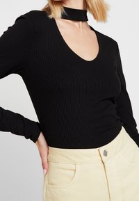 Musta ribbed pitkähiha TOPPI, jossa on syvä V-pääntie ja choker-mallinen kaula-aukko, yhdistettynä vaaleancremearvaiseen hameeseen, jossa on etutaskut.