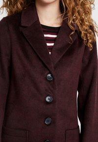 Manteau en mélange de laine bordeaux avec un design à boutonnage simple, un col cranté et six boutons noirs. Il présente une poche latérale et une texture douce.