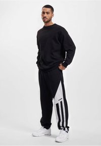 Schwarzer Hoodie kombiniert mit schwarzen Jogginghosen, die ein weißes diagonales Paneel und seitliche Streifen aufweisen, ergänzt durch weiße Sneaker.
