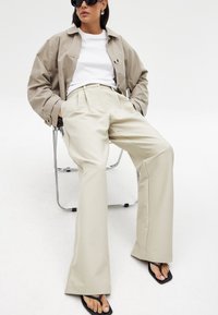 Bēša platā kāju bikses kopā ar baltu T-kreklu un gaiši bēšu jaku; modele sēž uz hroma krēsla, valkājot melnus saulesbrilles un sandales.